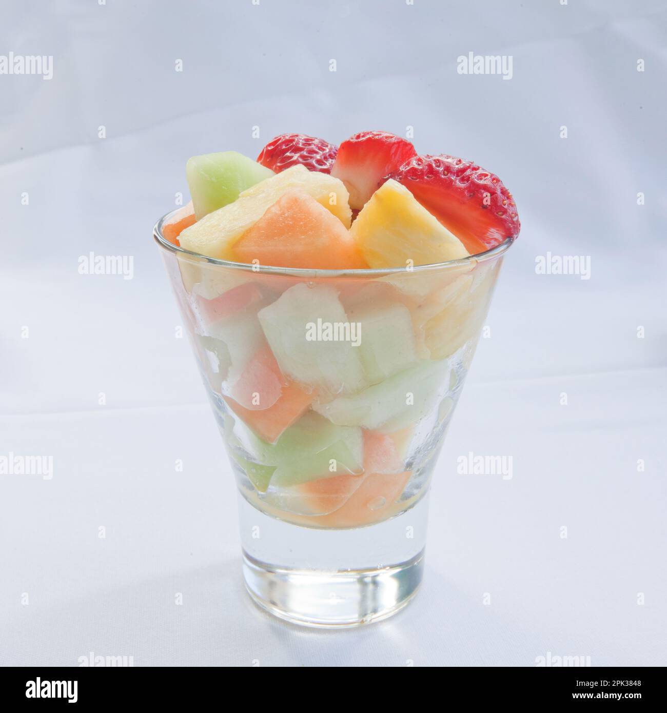 Frischer Obstsalat mit Erdbeeren, Melonen aus Cantaloupe und Honigtau sowie Ananasstücke in einem durchsichtigen Glasbecher auf weißem Leinenhintergrund Stockfoto