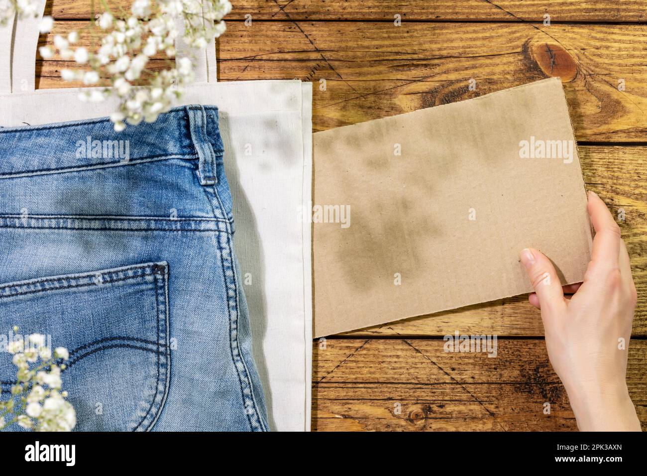 Nachhaltiges Stillleben mit Jeans auf einer Textiltasche und weiblicher Hand, die leere Pappe für deinen Text hält. Second-Hand-Shop. Kreisförmige Mode, umweltfreundlicher Freund Stockfoto