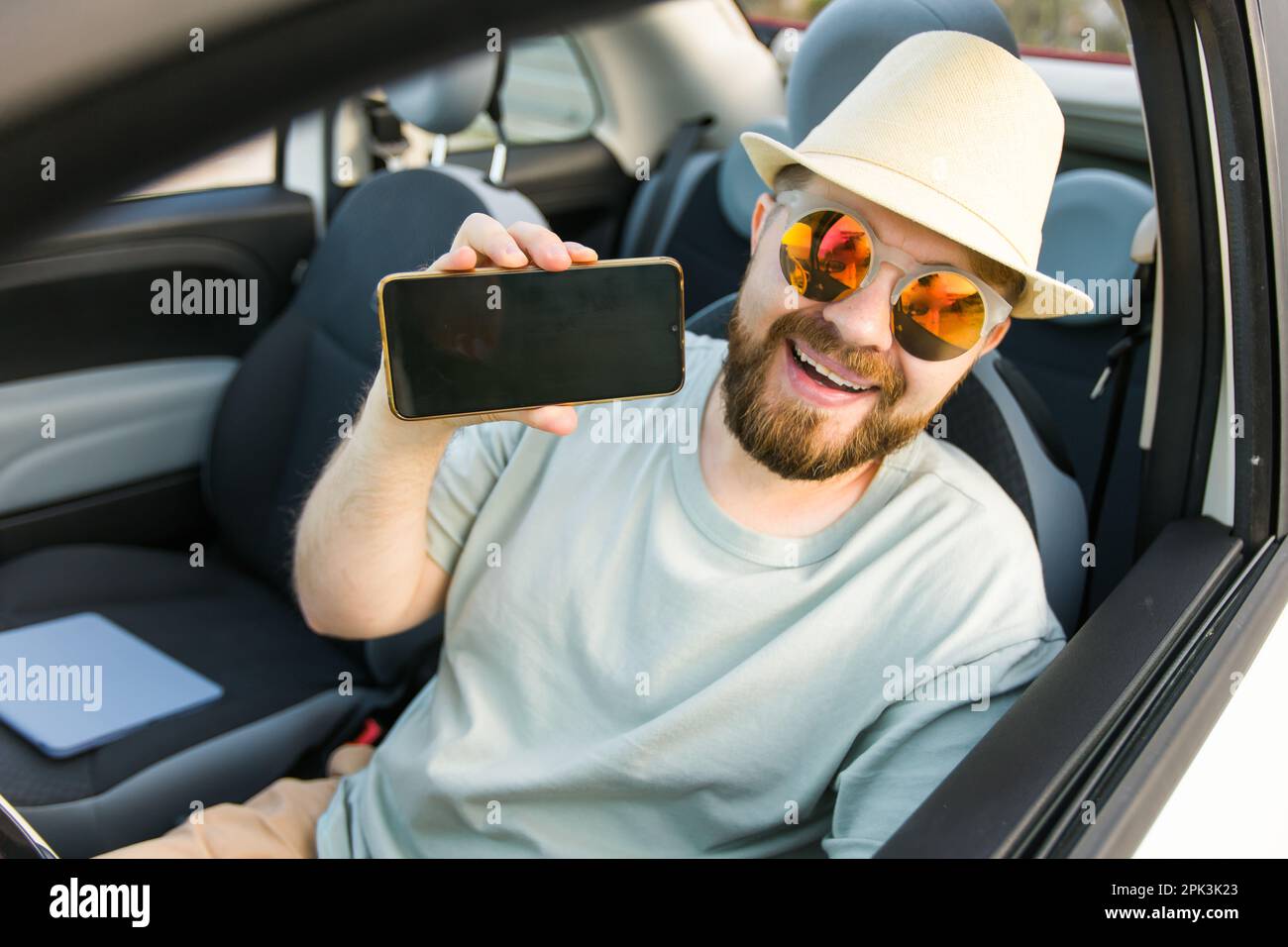 Lächelnder bärtiger Mann in Sonnenbrille zeigt Handy mit leerem Bildschirm für Modell, sitzt auf dem Fahrersitz in einem neuen Luxusauto-Cabriolet und hält gadge Stockfoto