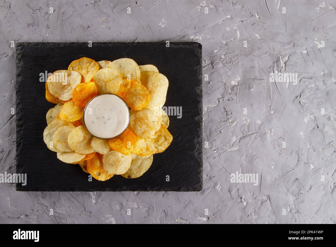Draufsicht auf einen Haufen Kartoffelchips mit cremeweißer Dippsauce auf schwarzem Schiefertafel auf einem grauen Putztisch. Klassische Auswahl an Snacks und Dips Stockfoto