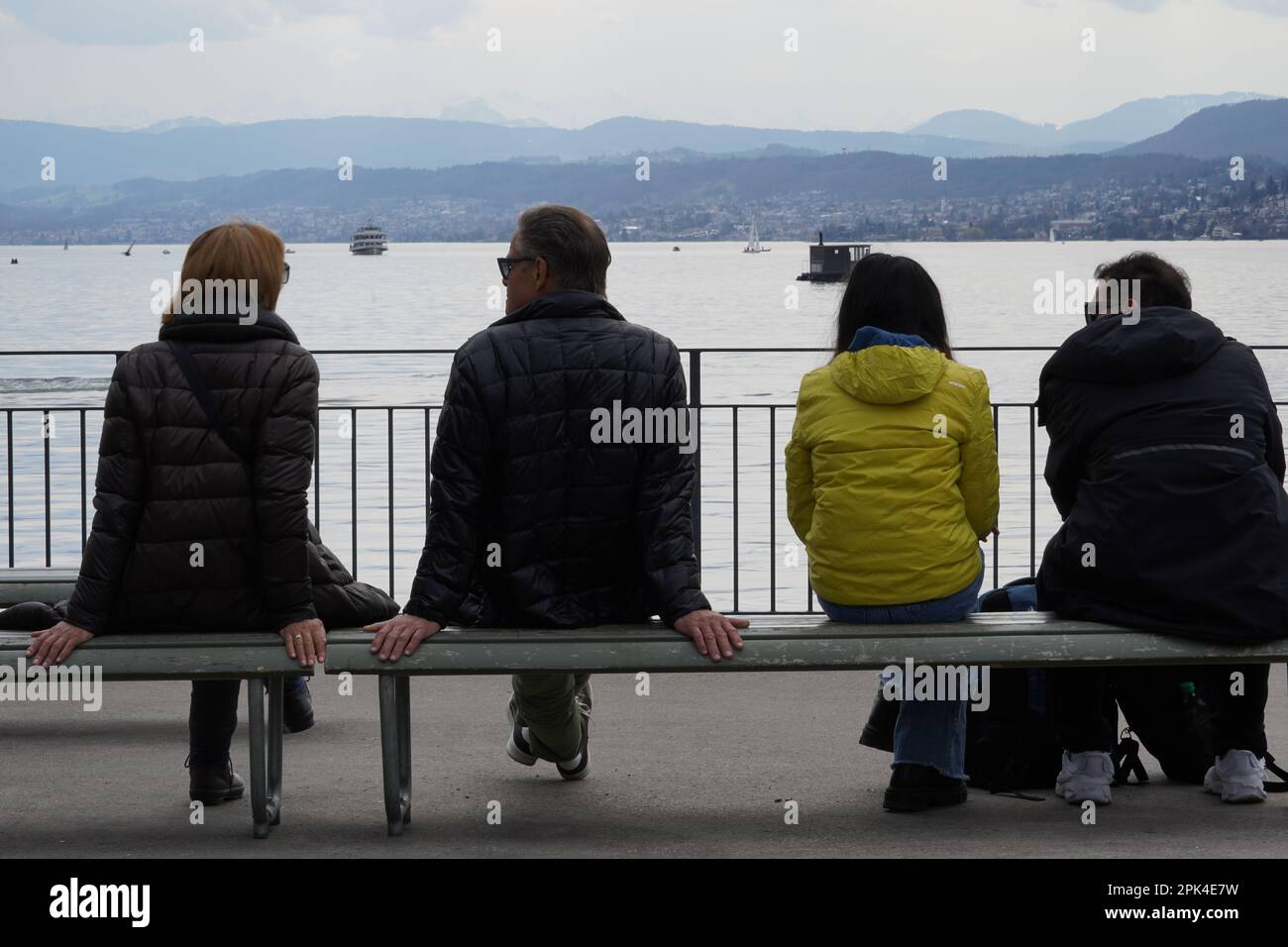 Zwei heterosexuelle Paare, die auf einer Bank am Ufer des Zürichsees sitzen. Rückansicht mit See und Schweizer Alpen im Hintergrund. Stockfoto