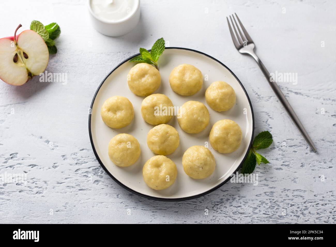 Glutenfreie gedämpfte, faule Teigtaschen mit Apfelfüllung auf einem weißen Teller, dekoriert mit Minze, umgeben von einem halben Apfel und saurer Sahne auf einem Stockfoto