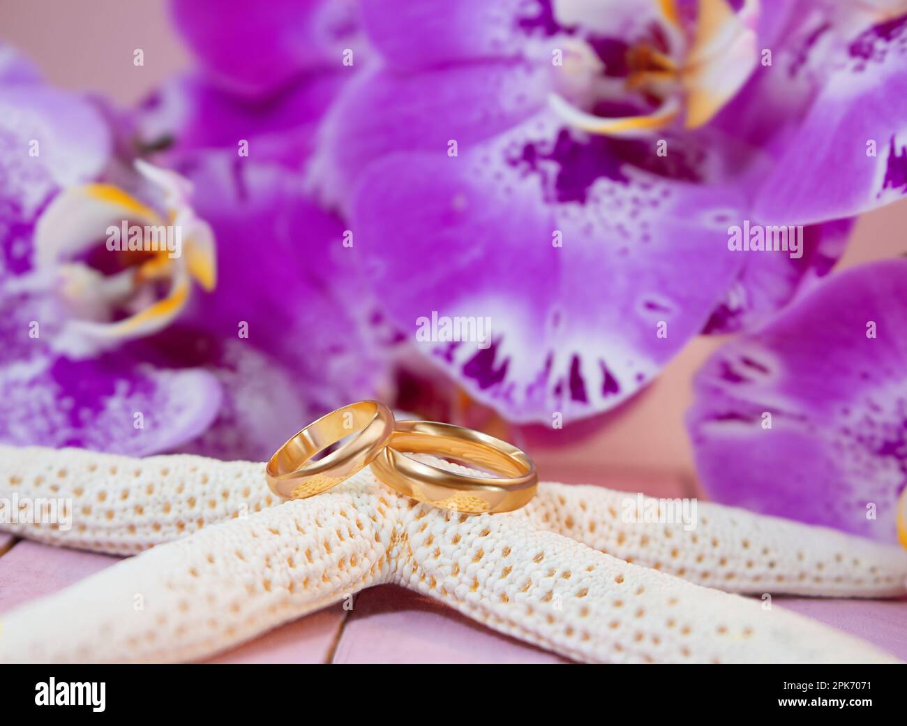 Goldene Eheringe auf weißen Seesternen hinter lila Orchideen auf pinkfarbenem Holzbrett. Feiern, Zeremonien, Flitterwochen Stockfoto