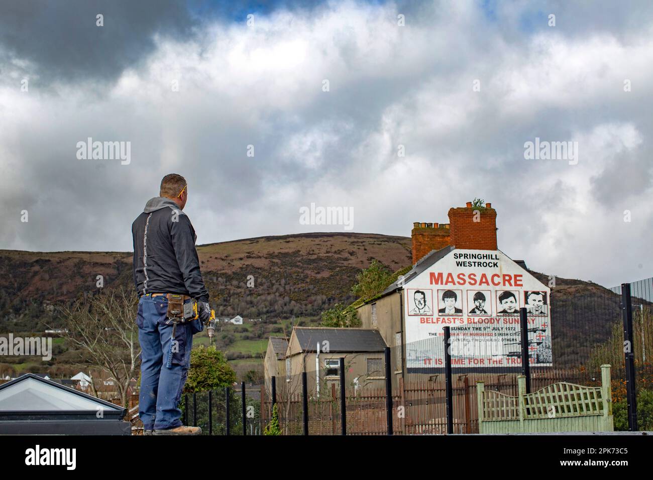 Dachdeckerbauer mit Handbohrmaschine, die auf das Wandgemälde in West Belfast blickt, zum Gedenken an das Springhill WestRock Massaker, 9. Juli 1972, Belfast. Stockfoto