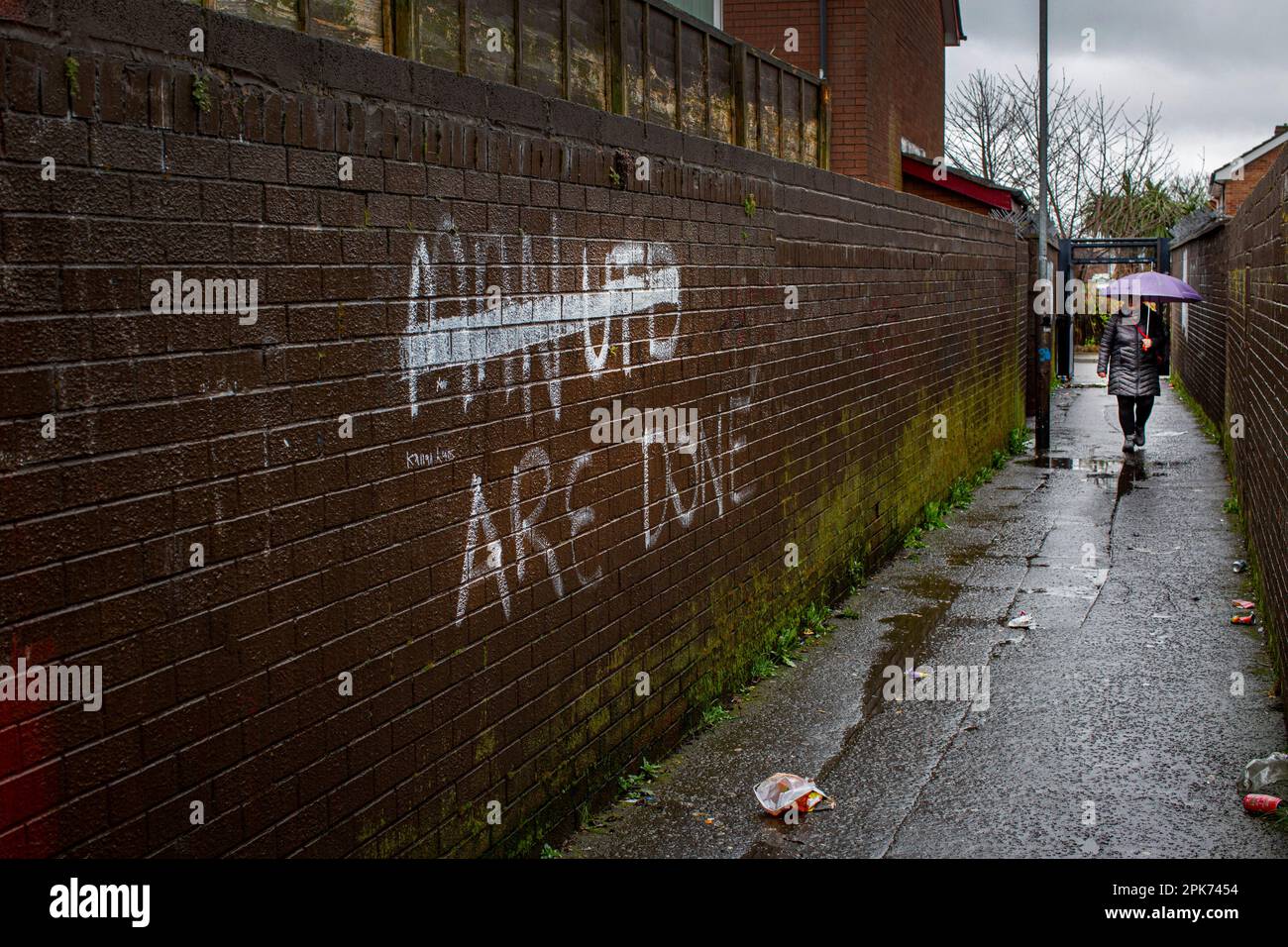 Eine Frau mit Schirm überquert paramilitärisches Graffiti von Loyalist UDA/UFF an einer Wand in West Belfast. Stockfoto