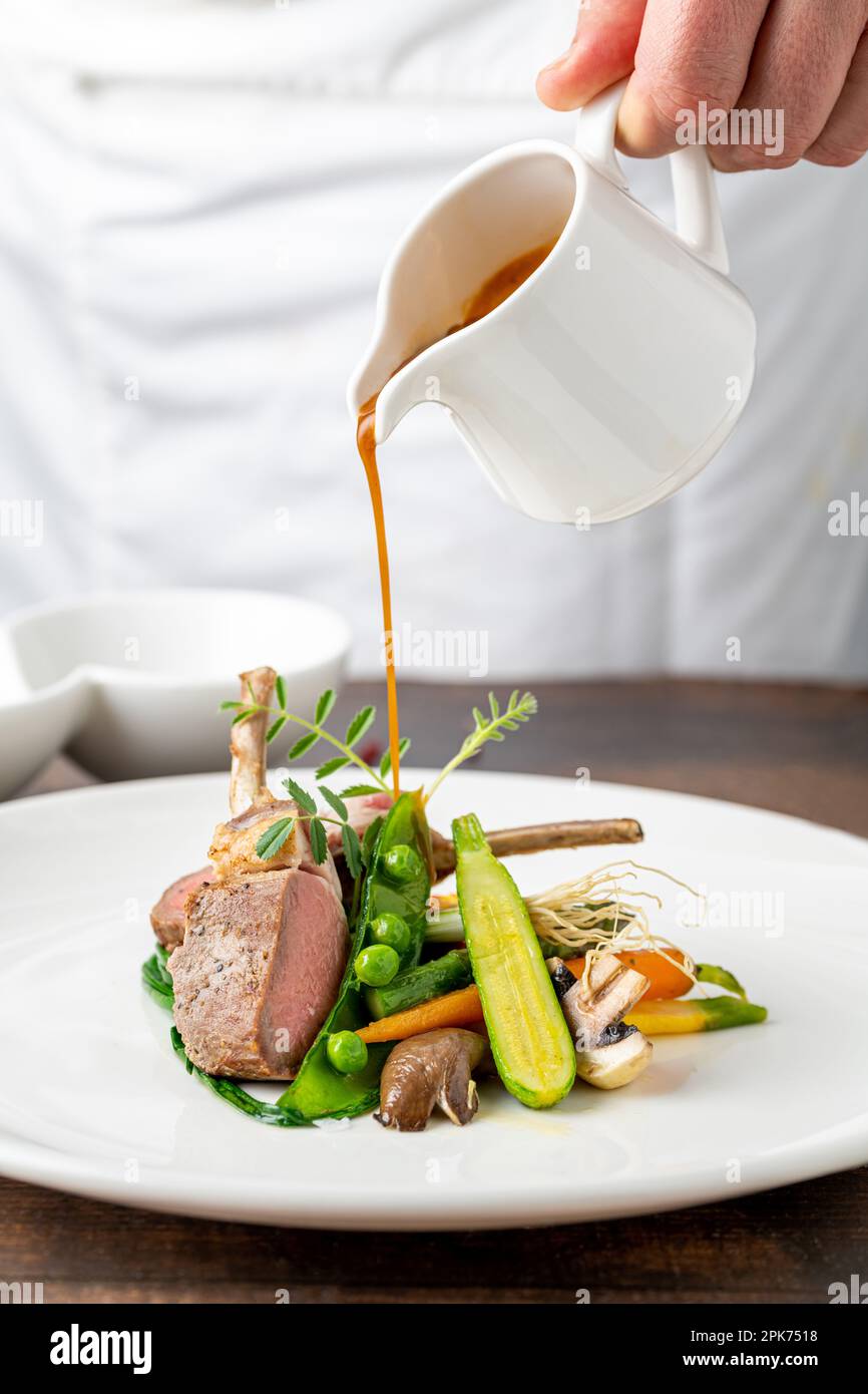 Der Koch gießt in einem feinen Restaurant Sauce auf Lammkoteletts mit verschiedenen Gemüsesorten Stockfoto