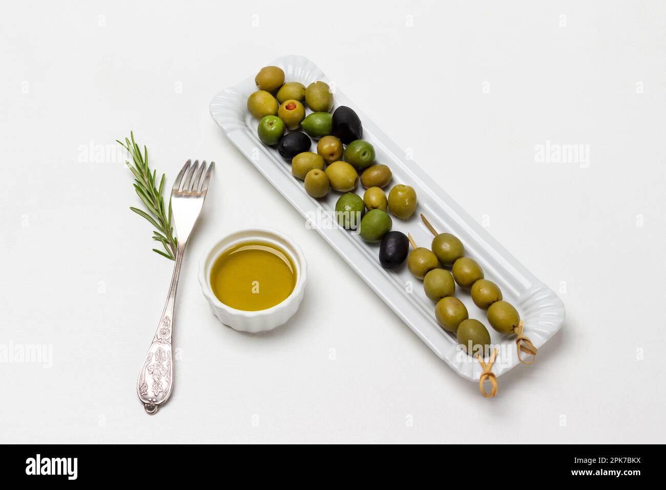 Grüne Oliven auf Spießen. Oliven auf weißem Teller. Olivenöl in der Schüssel. Gabel auf Tisch. Weißer Hintergrund. Stockfoto