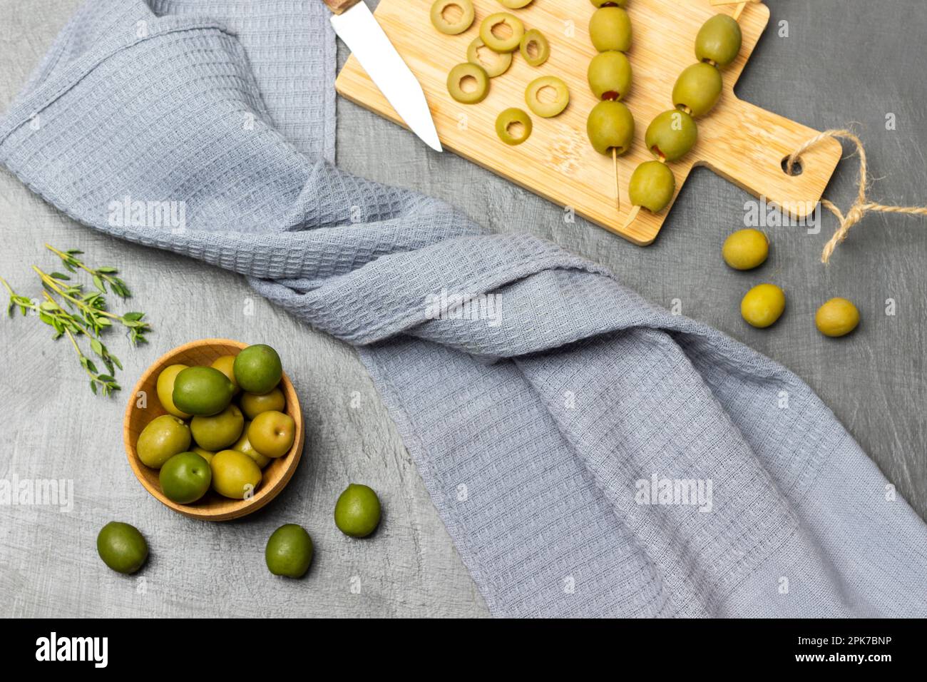 Grüne Oliven in einer Holzschüssel. Geschnittene Oliven und Messer auf dem Schneidebrett. Graue Serviette auf grauem Hintergrund. Flach verlegt. Stockfoto