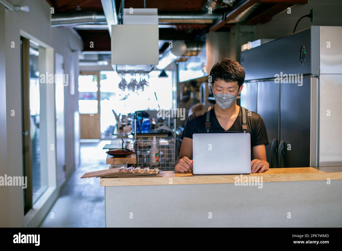 Ein Mann mit einer Gesichtsmaske in einer Restaurantküche, der einen Laptop benutzt, der Besitzer oder Manager. Stockfoto