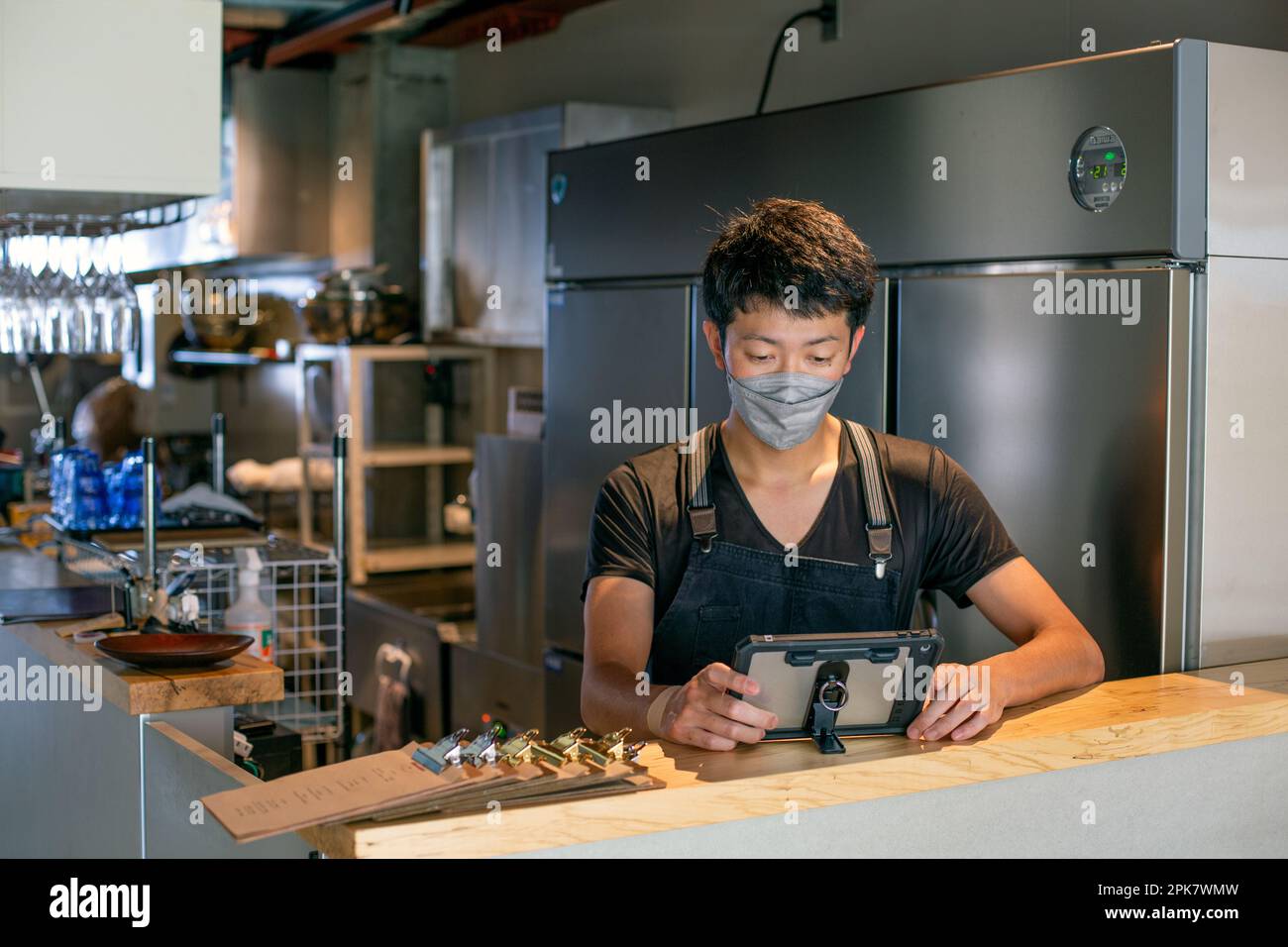 Ein Mann, der eine Gesichtsmaske an der Theke einer Restaurantküche trägt, ein digitales Tablet benutzt, der Besitzer oder Manager. Stockfoto
