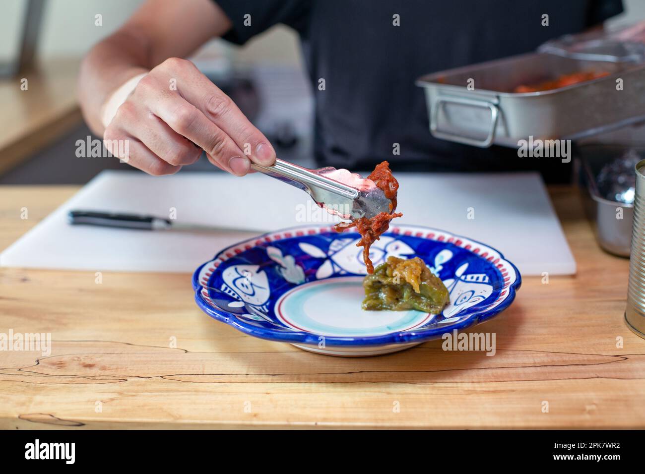 Ein Mann, der Essen in einer Restaurantküche zubereitet, indem er mit einer Zange Gemüse auf einen Teller stellt. Schließen. Stockfoto
