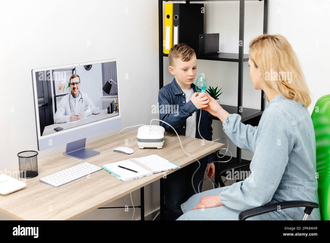 Kaukasischer kleiner Junge, der zu Hause mit einem Vernebler inhaliert. Eine junge Mutter, die ihrem Sohn beim Einatmen mit einem Vernebler hilft, bei einem Online-Meeting mit einem D. Stockfoto