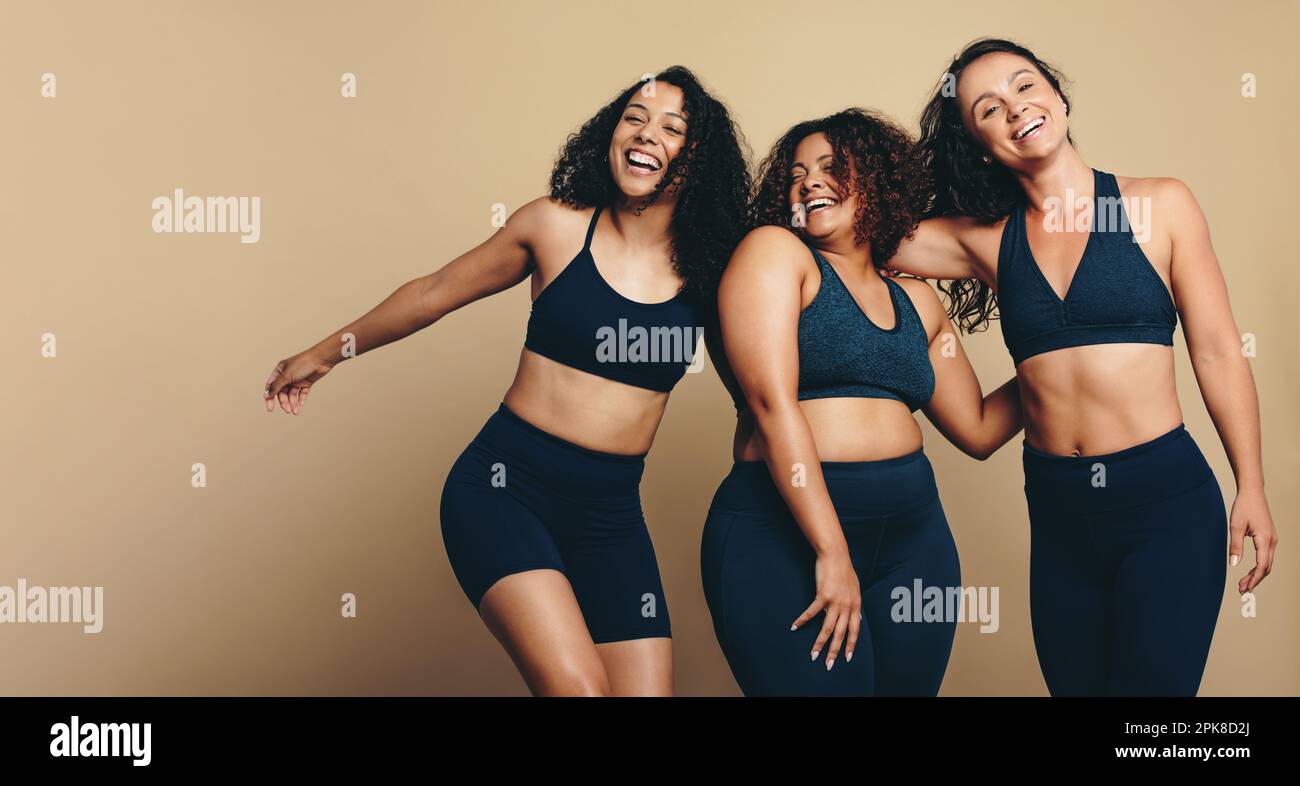Drei junge Erwachsene Frauen, die vor Glück und Zusammensein in einem Sportstudio lächeln. Sie tragen Sportbekleidung und stylische Frisuren und feiern damit ihren eigenen Look Stockfoto