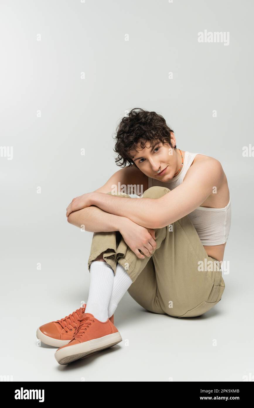 Junger und stilvoller, großer Mensch, der sich in die Knie drückt und dabei auf grauem Hintergrund sitzt, Stockbild Stockfoto