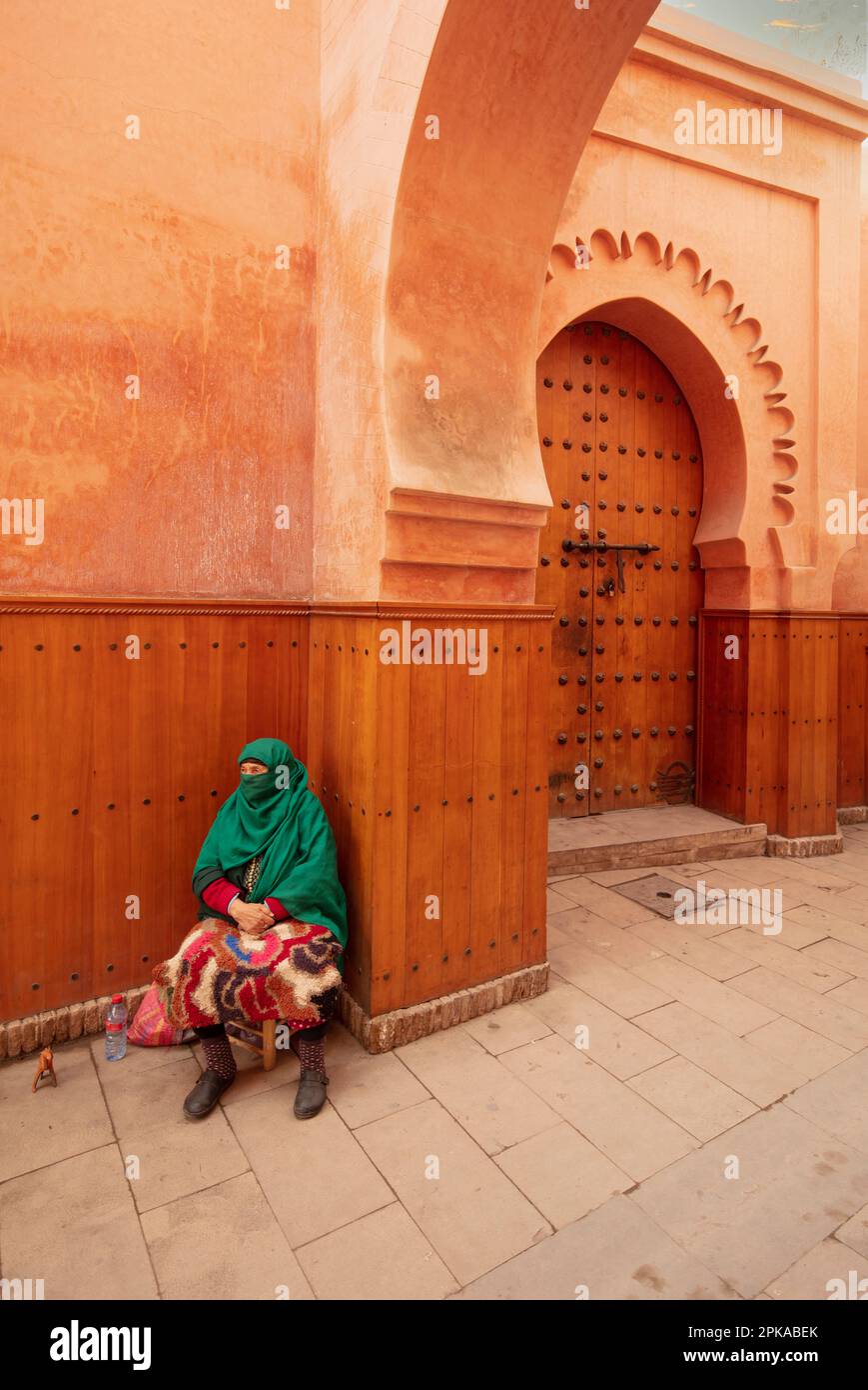Marokko, Marrakesch, Madersa Ben Youssef Madrasa, schöne Architektur, sitzende Frau mit grünem Schleier vor dem braunen orangefarbenen Gebäude Stockfoto