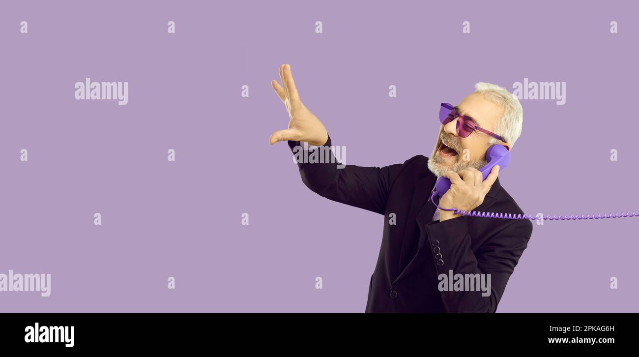 Fröhlicher und stilvoller Seniorenmann, der auf einem in Pastelllila gehaltenen Festnetztelefon telefoniert. Stockfoto