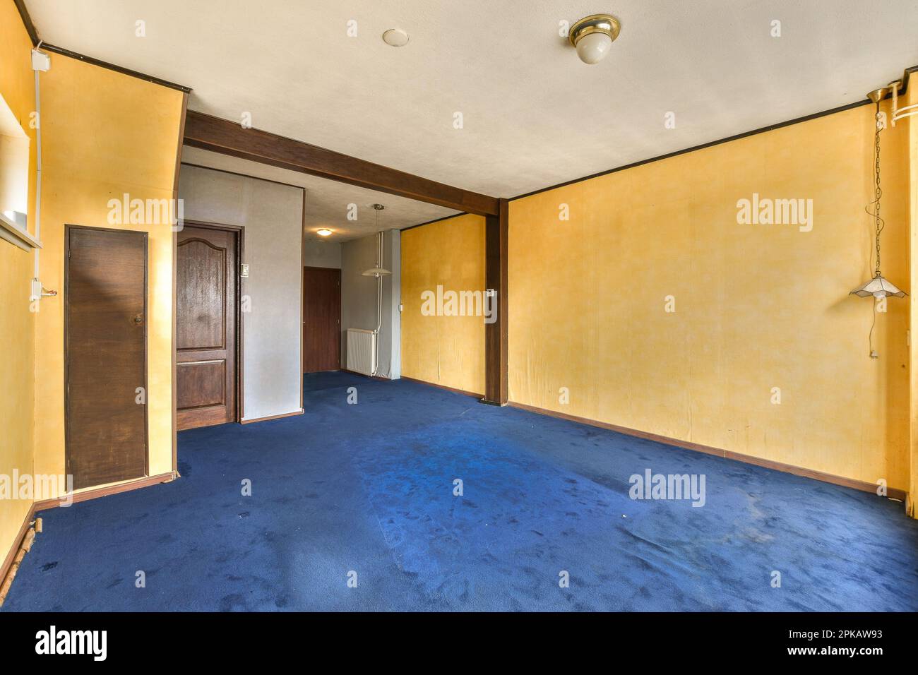 Ein leeres Zimmer mit gelben Wänden und blauem Teppich in der Mitte des Zimmers, es gibt eine Tür, die zu einem anderen Zimmer führt Stockfoto