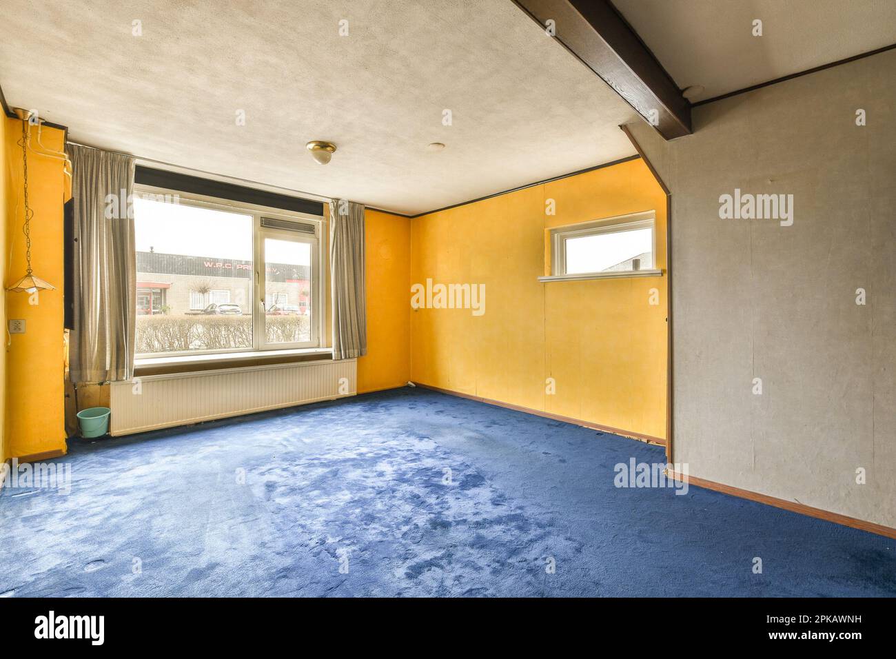 Ein leeres Zimmer mit gelben Wänden und blauem Teppichboden vor dem Fenster mit Blick auf die Straße Stockfoto