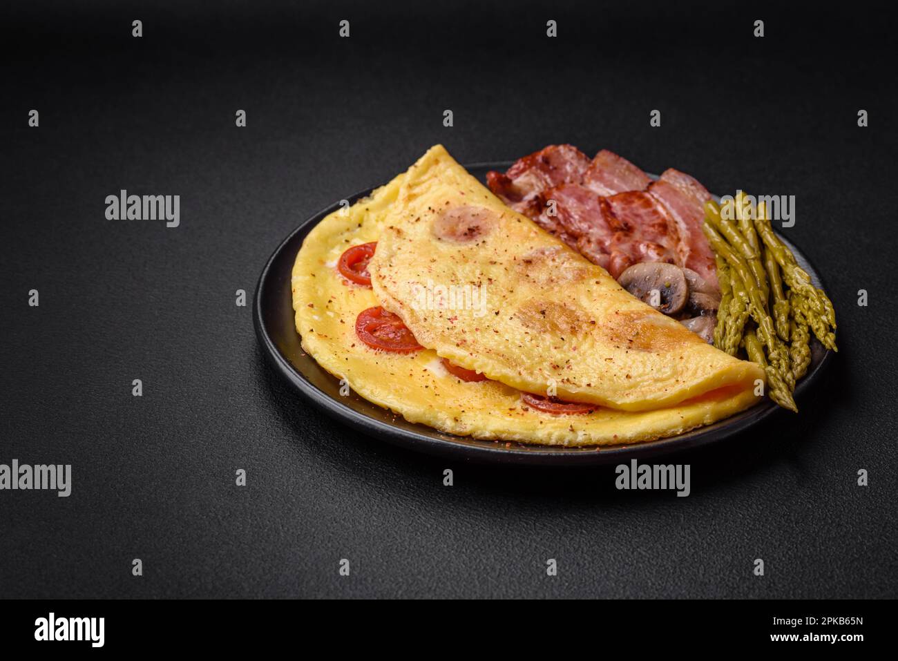 Köstliches frisches Omelett mit Kirschtomaten, Speck, Spargel und Gewürzen auf dunklem Betonhintergrund Stockfoto