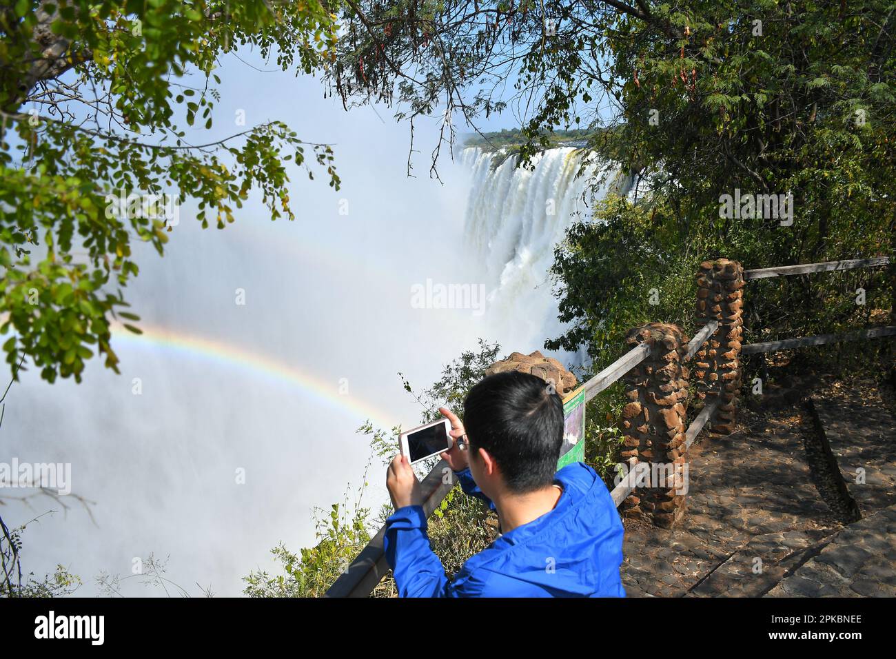 Ein Tourist macht Fotos vom Victoria-Wasserfall an der Grenze zwischen Sambia und Simbabwe. Die mächtigen Victoria Falls am Sambesi River sind einer der meistbesuchten touristischen Orte in Afrika. Victoria Falls liegt direkt an der Grenze zwischen Sambia und Simbabwe im südlichen Afrika. Es ist einer der höchsten Wasserfälle der Welt. Stockfoto
