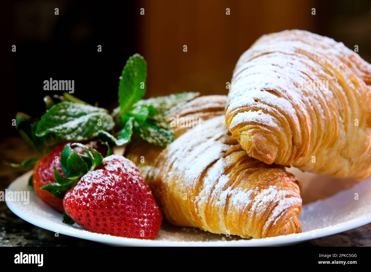 Puderzucker, der auf ein Croissant auf einem weißen Teller gestreut wird ein Croissant mit Erdbeeren zwei reife Erdbeeren sind mit Puderzucker-Minze gestreut, hinterlässt rote, transparente Sauce Stockfoto