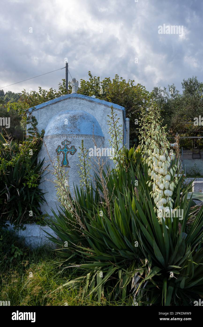 Blaue Kapelle mit Kreuz, in einem Garten an der Küste. Weißer Engel auf dem Dach. Blühender Yucca. Blauer Himmel mit weißen Wolken. Kinira, Thassos (Tassos) isla Stockfoto