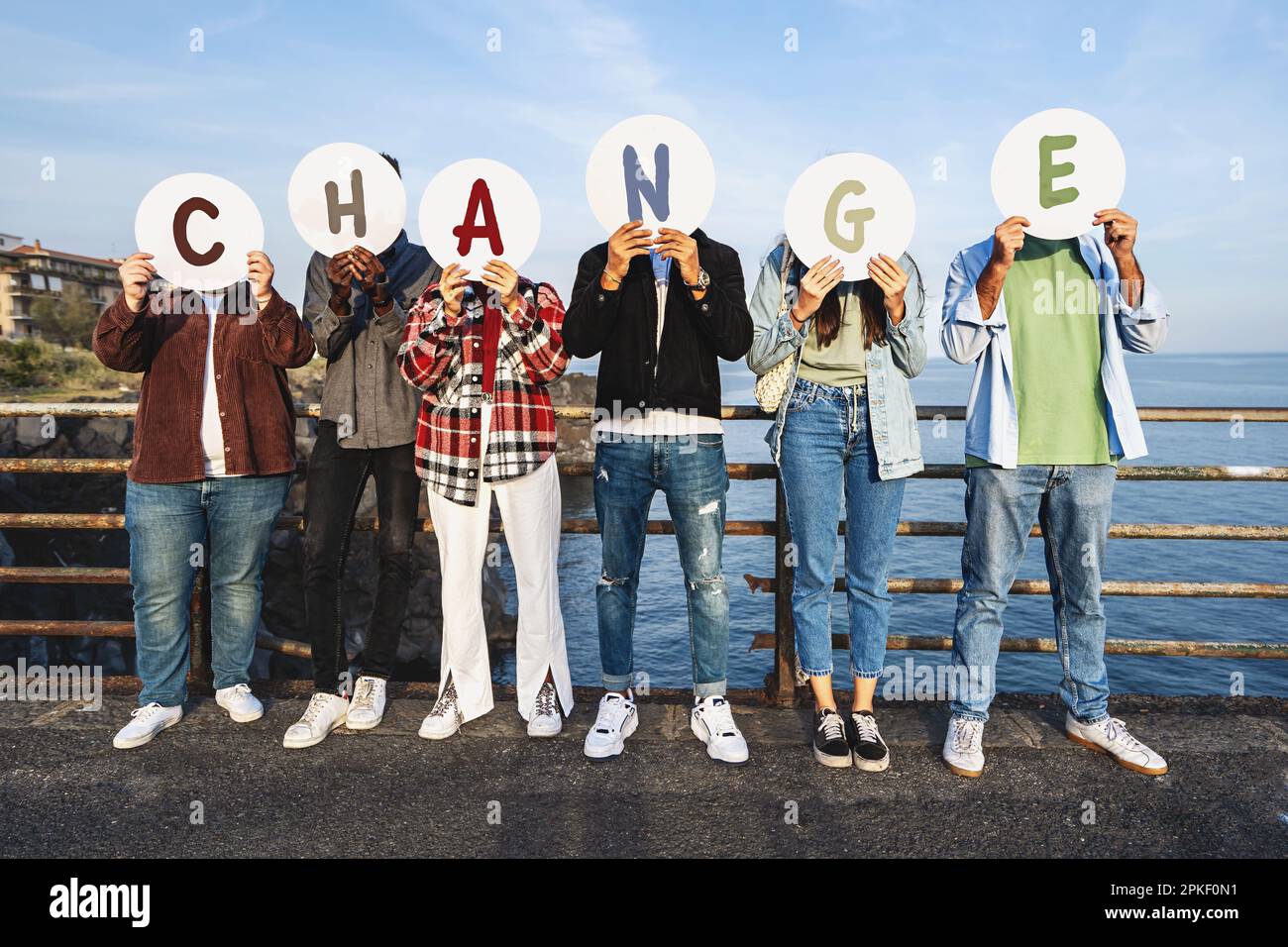 Eine multiethnische Gruppe junger Menschen, die ihre Gesichter mit Pappscheiben bedecken, vor einem Geländer stehen, mit dem Meer und Felsen dahinter - das Stockfoto