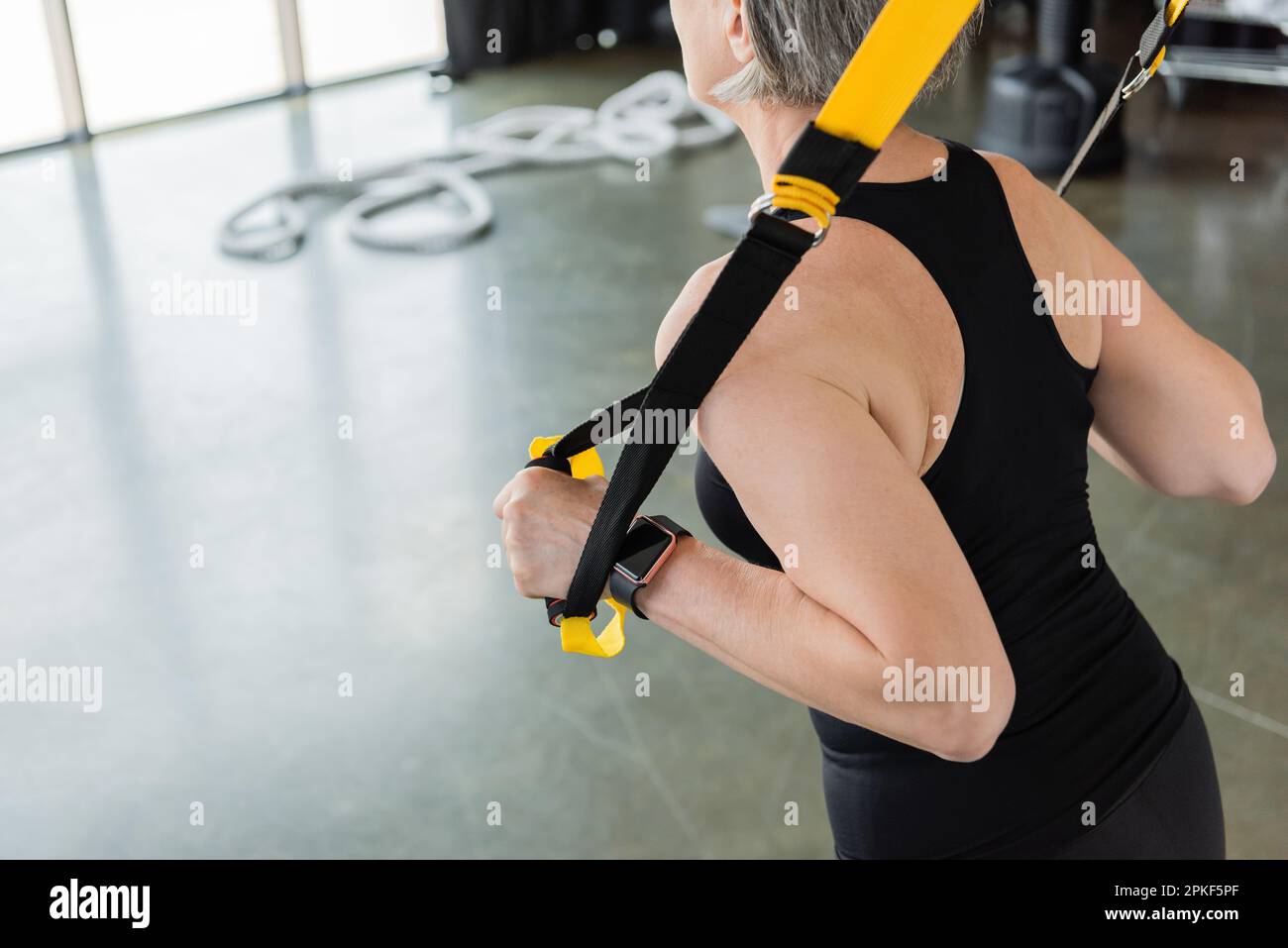 Verkürzte Ansicht einer Seniorin in schwarzer Sportbekleidung, die im Fitnessstudio mit Aufhängebändern trainiert, Stockbild Stockfoto