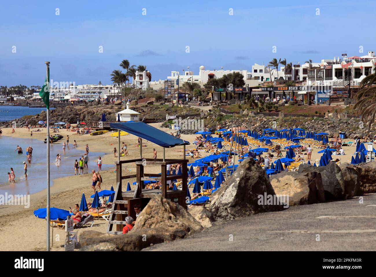 Weg an einer geschäftigen Strandszene, Playa Blanca, Lanzarote mit hölzerner Plattform Rettungsschwimmerstation. Februar/März 2023. Stockfoto