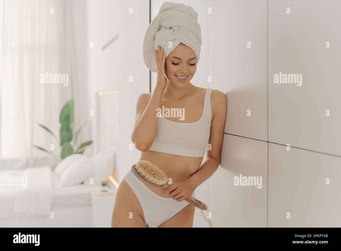 Die entspannte junge Weiße trägt ein Handtuch auf dem Kopf, fühlt sich nach dem Duschen und der Trockenmassage mit Bürste erfrischt an, posiert im gemütlichen Badezimmer im Stockfoto