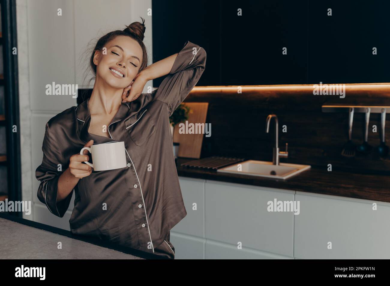 Eine schöne, entspannte junge Frau in einem gemütlichen Seidenpyjama, die sich aus dem Schlaf ausstreckt und früh am Morgen mit einer Tasse Kaffee in der Hand aufwacht, während sie steht Stockfoto