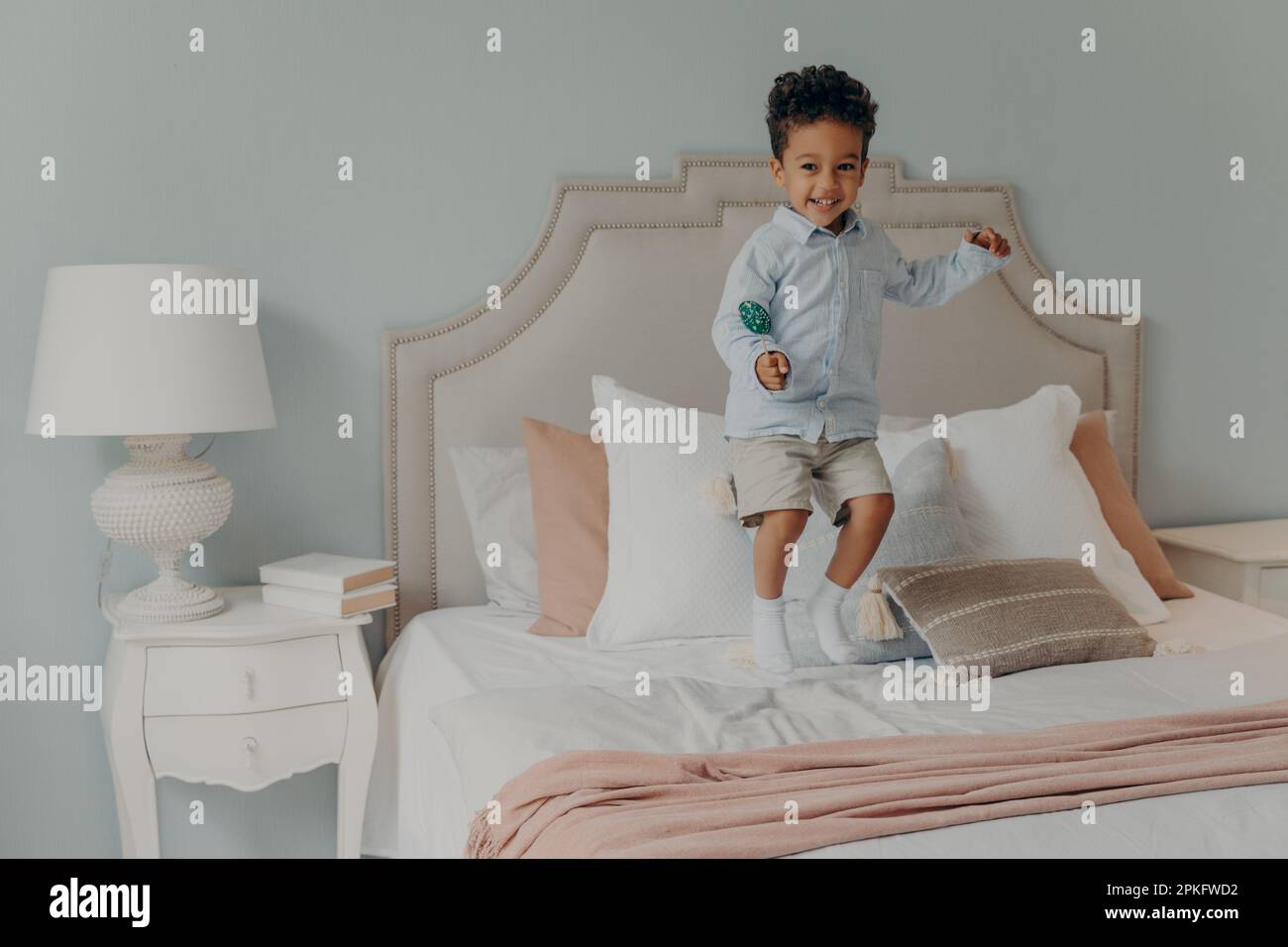 Ein glücklicher, kleiner, energetischer afroamerikanischer Junge, der allein auf dem Bett seiner Eltern sprang, während er köstlichen Lollipop hielt, Süßigkeiten aß, Chaos verursachte, während nein Stockfoto