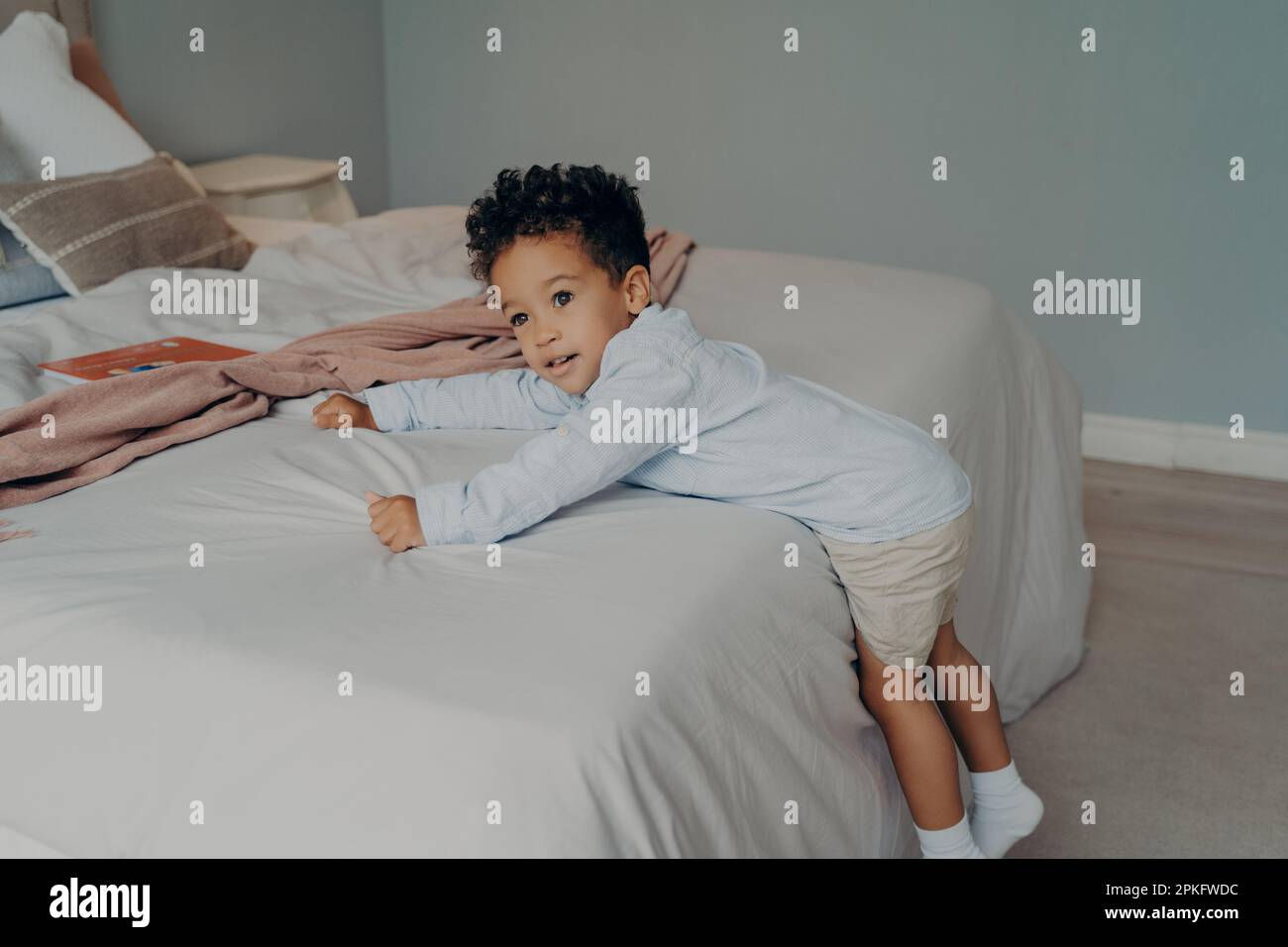 Süßer kleiner afro-amerikanischer Junge in stilvollen Baumwollkleidung, der versucht, auf ein weißes, weiches Bett zu klettern, sich am Bettlaken festzuhalten, während er in einem hellen Bett spielt Stockfoto