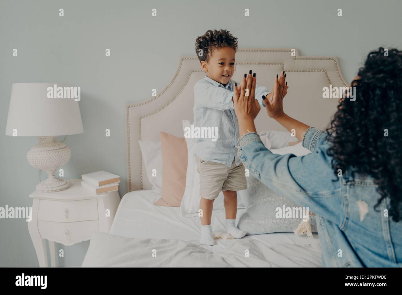 Süßes kleines afro-amerikanisches Kind spielt spannende Spiele auf einem großen Bett in einem hellen Schlafzimmer mit seiner fürsorglichen lockigen Mutter, die ihre Hände zusammenhält. Werteliste Stockfoto