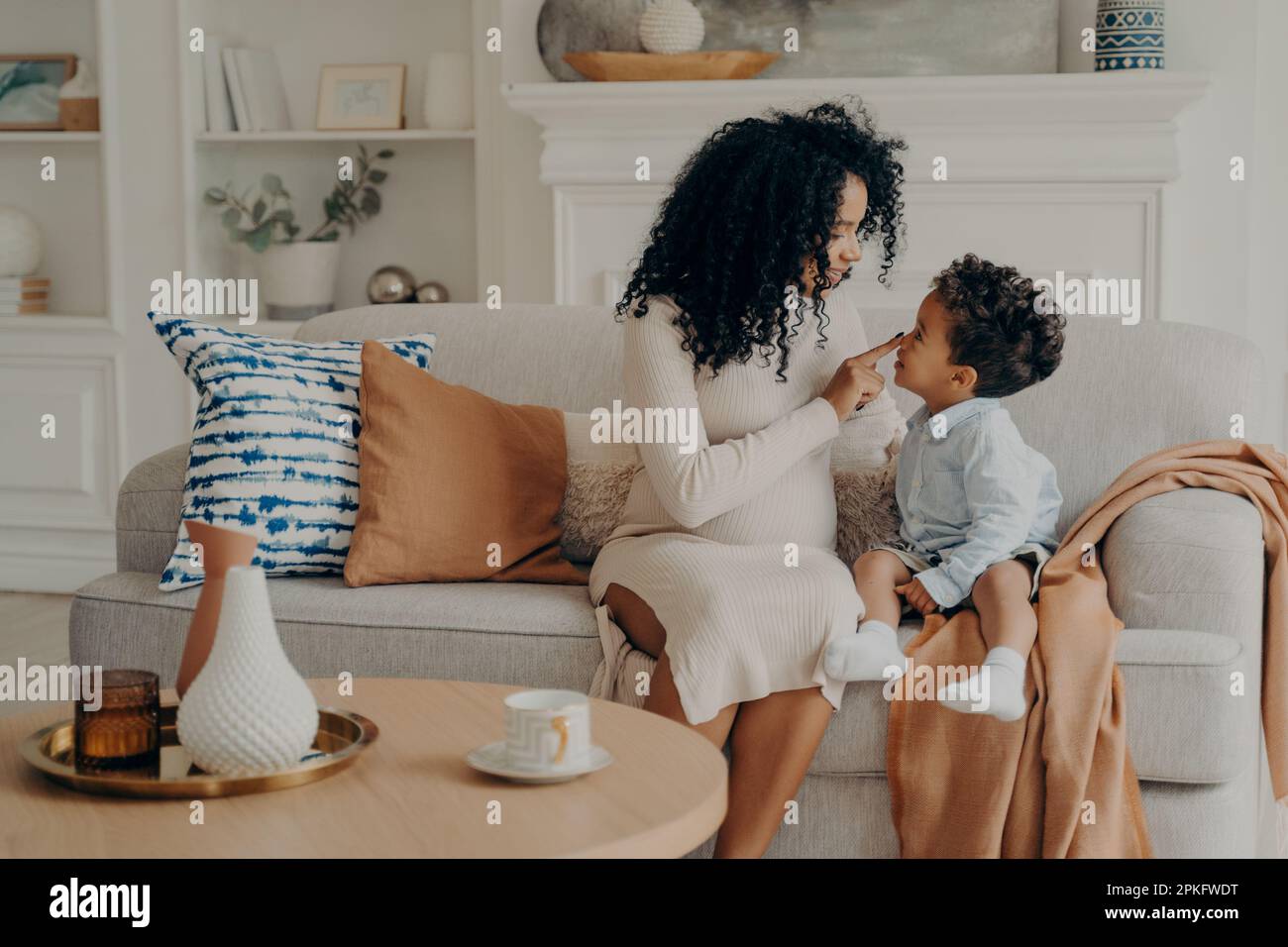 Schwangere Mutter und Sohn, afro ethnische Rasse, sitzen auf dem Sofa im modernen Wohnzimmer, mom redet mit dem Jungen, der sich mit der Fingerspitze seiner Nase berührt, Kiefer Stockfoto