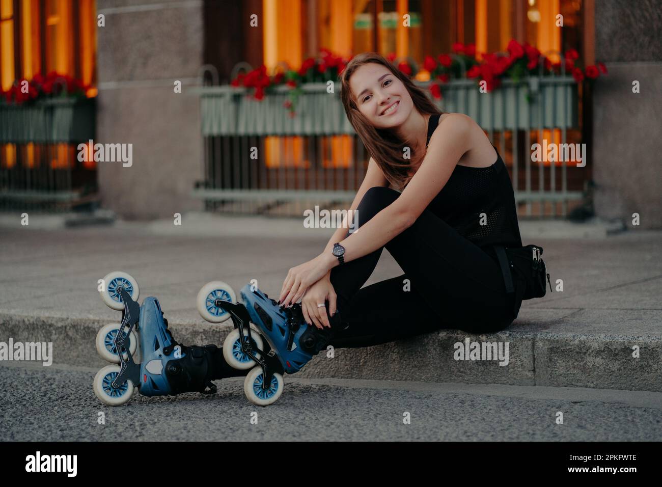 Eine attraktive junge europäische Frau trägt Rollerskates und macht nach dem Fahren eine Pause. Sie trägt schwarze Aktivbekleidung und hat ein fröhliches Lächeln im Gesicht. P Stockfoto