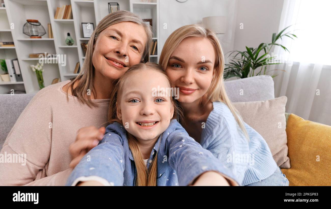 Glückliche Großmutter, Mutter und Enkelin sitzen auf dem Sofa im Wohnzimmer, machen Selfies, machen einen Videoanruf mit Verwandten, winken Stockfoto