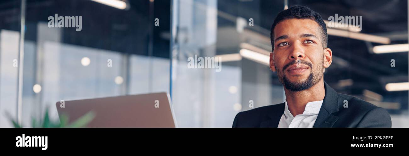 Schöner afrikanischer Geschäftsmann, der in einem modernen Büro sitzt und mit einem Laptop arbeitet und wegschaut Stockfoto