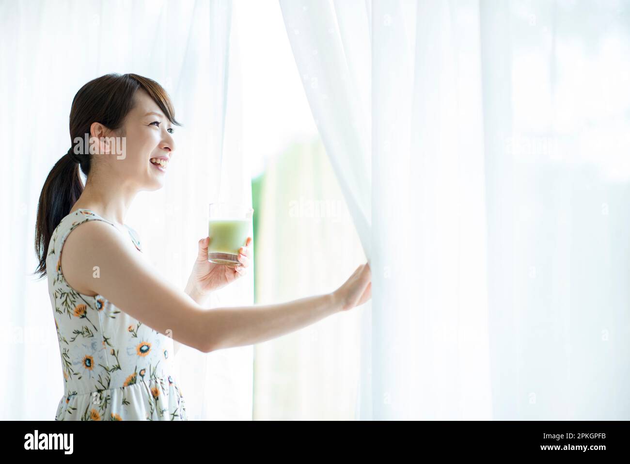 Eine Frau, die einen Smoothie hält und Vorhänge öffnet Stockfoto