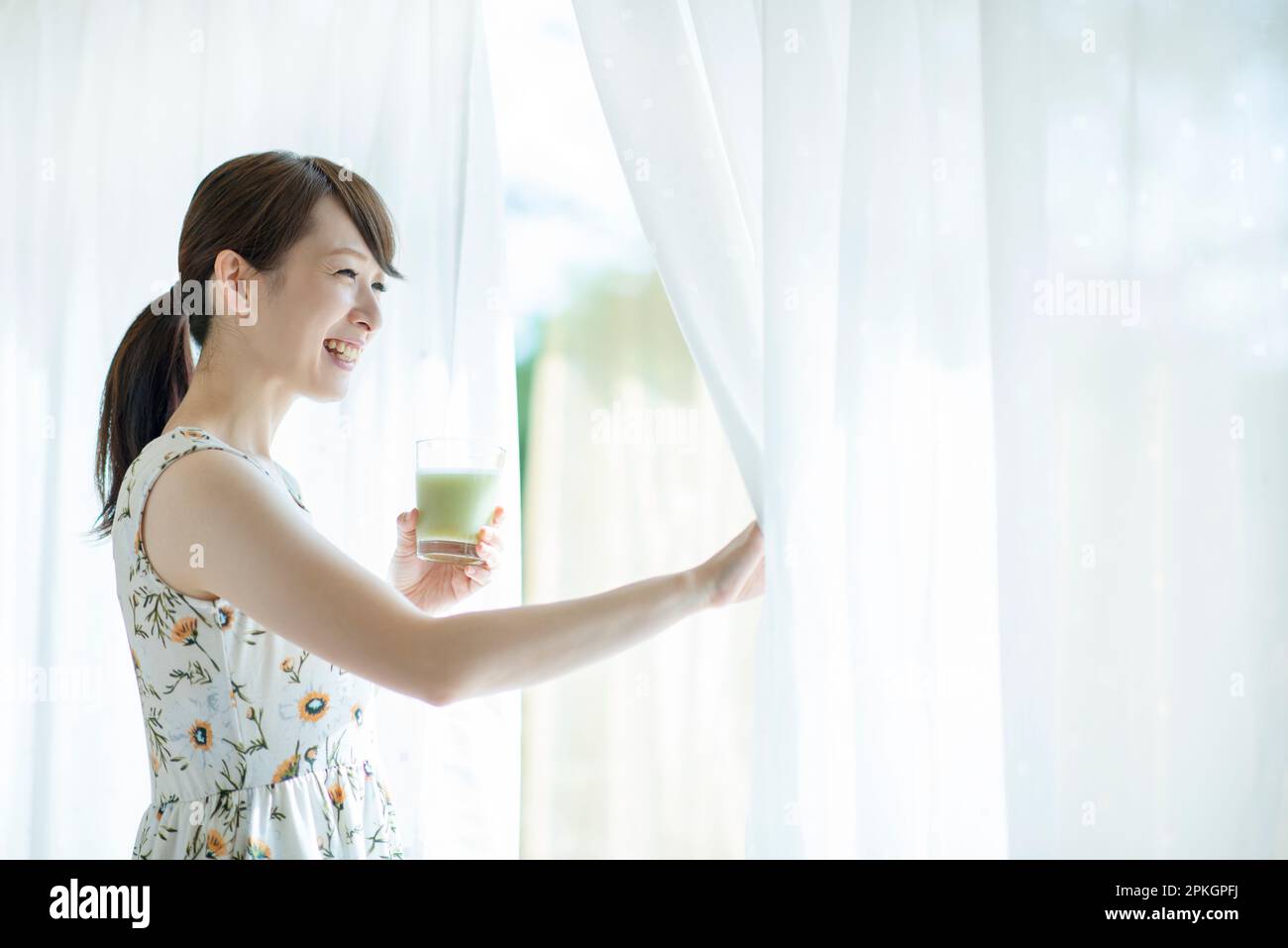 Eine Frau hält einen Smoothie und öffnet die Vorhänge Stockfoto