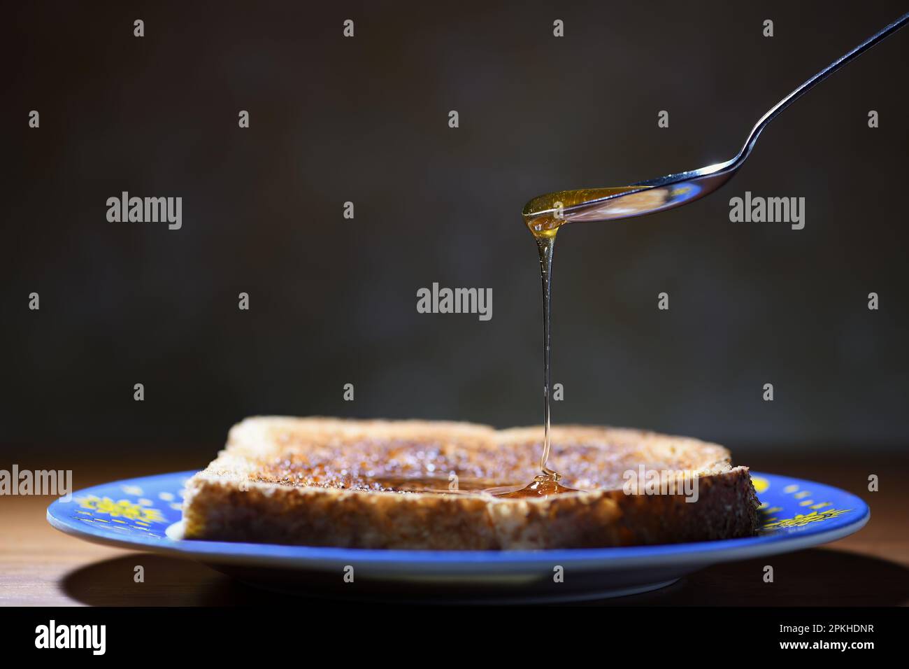 Eine Seitenansicht eines geneigten Löffels, der frischen Honig auf eine getoastete Scheibe Brot tropft, auf einem Teller, in sanfter, stimmungsvoller Beleuchtung mit Kopierbereich auf der linken Seite Stockfoto