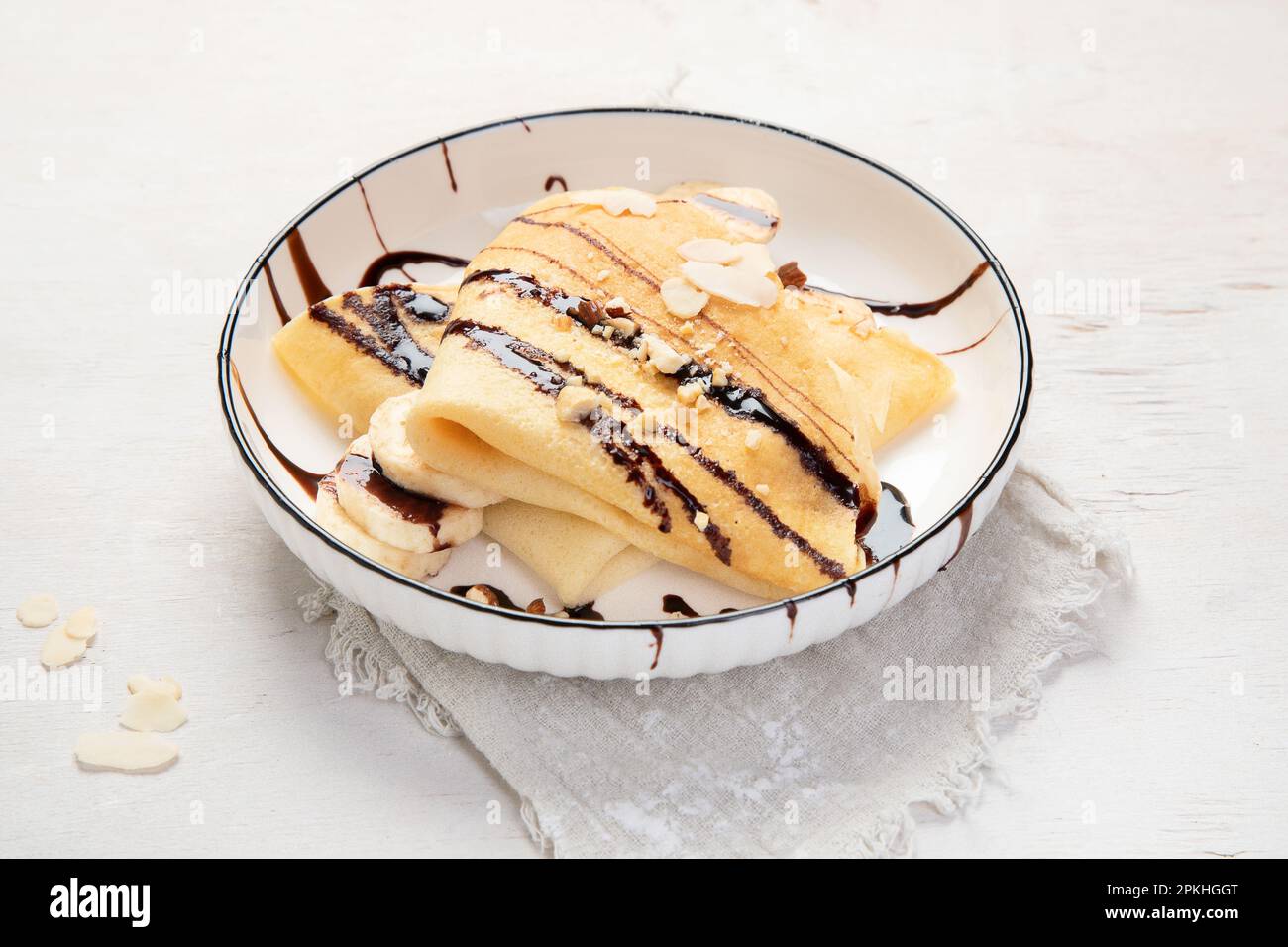 Crepe mit Banane, gehackten Mandeln und Schokoladensoße auf weißem Teller, Blick vom Tisch auf weißem Hintergrund. Köstliche crepes nach französischer Art. Stockfoto