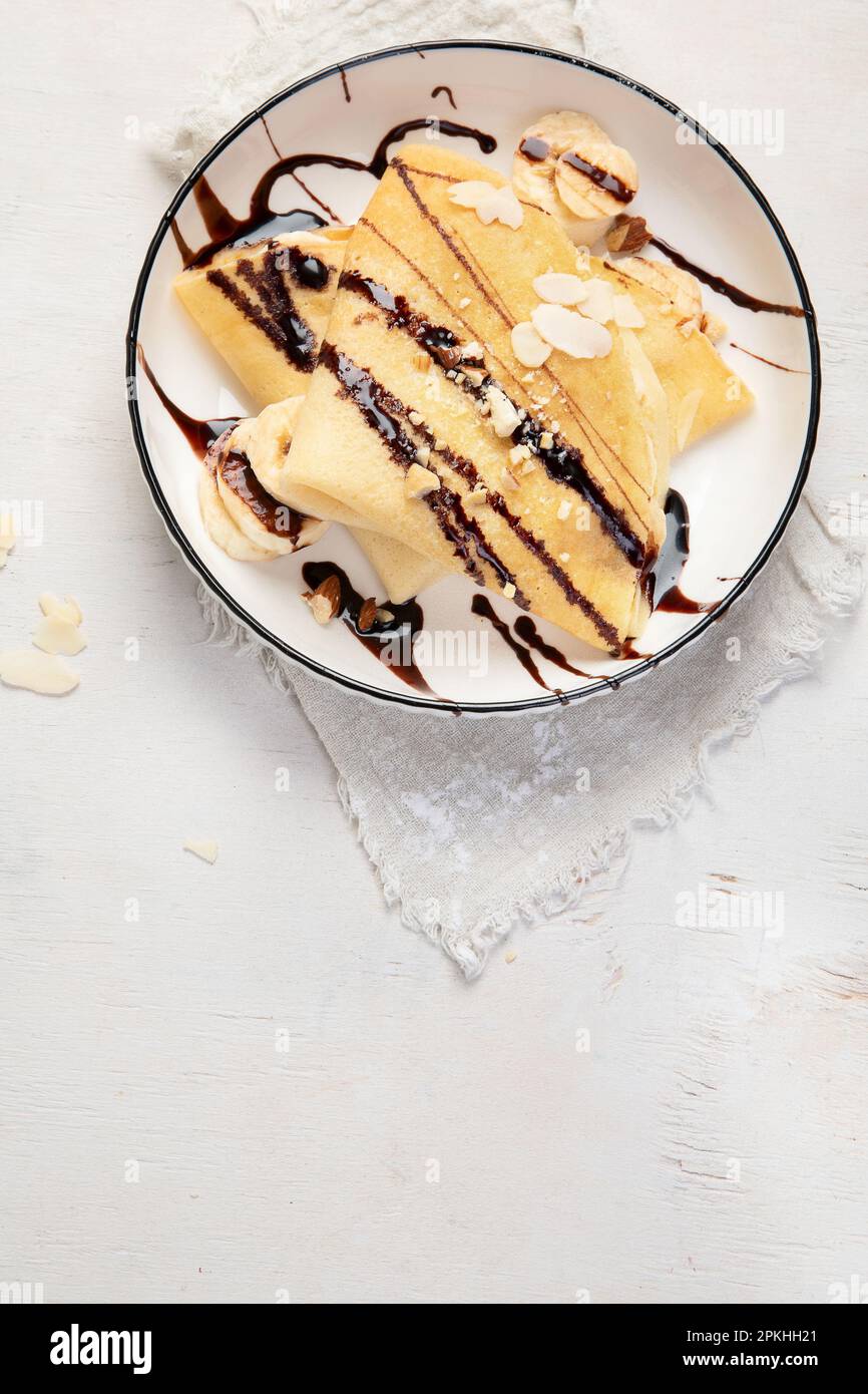 Crepe mit Banane, gehackten Mandeln und Schokoladensoße auf weißem Teller, Blick vom Tisch auf weißem Hintergrund. Köstliche crepes nach französischer Art. Stockfoto