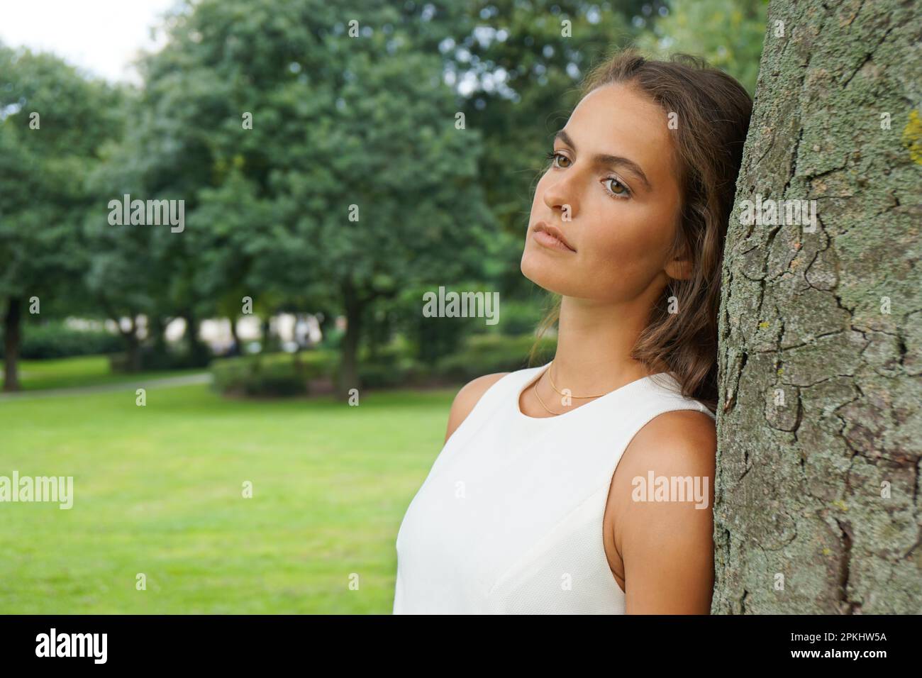 Traurige junge Frau, die sich gegen den Baum lehnt und abwesend blickt Stockfoto