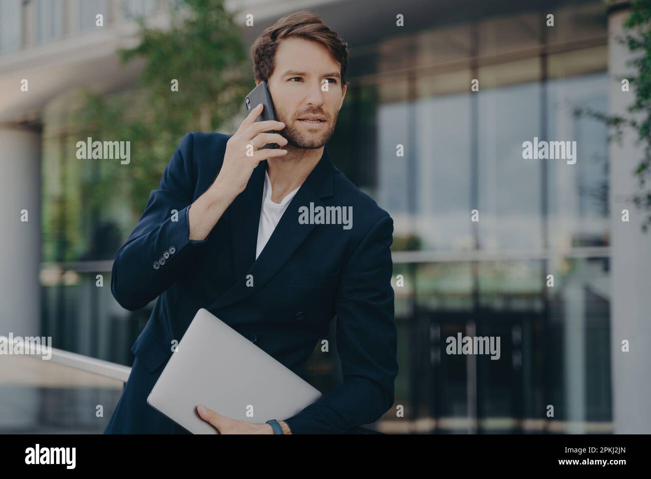 Ein intelligenter Geschäftsmann spricht telefonisch, während er zu Fuß ins Büro geht, wartet auf ein Geschäftstreffen, trägt formelle Kleidung und verwendet moderne Geräte für die Kommunikation Stockfoto