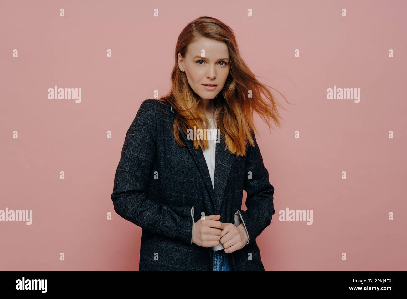 Mittelgroße Aufnahme einer attraktiven Frau mit Ingwerhaaren, die in die Kamera schaut. Sie trägt eine dunkle, formelle Jacke und ein weißes Oberteil, das isoliert über dem rosa Studiohintergrund steht Stockfoto