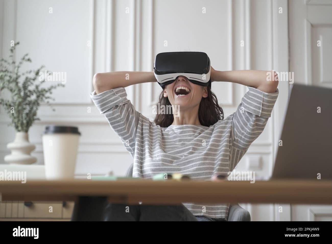 Erstaunte Büroangestellte in VR-Brillen, die mit Freude lachte und sich mit den Händen hinter dem Kopf in den Stuhl lehnte, während sie aus der Ferne vom Heimbüro aus arbeitete, che Stockfoto