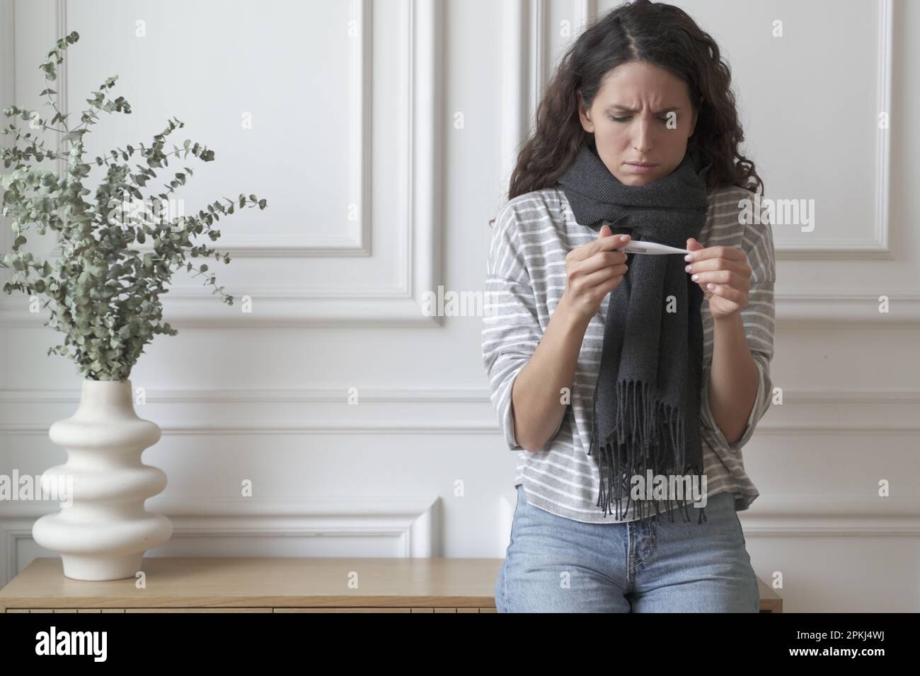 Ungesunde kranke junge Italienerin, die ein Thermometer hält und hohe Temperaturen misst, während sie zu Hause drinnen steht, unglückliche besorgte weibliche Gefühl Stockfoto