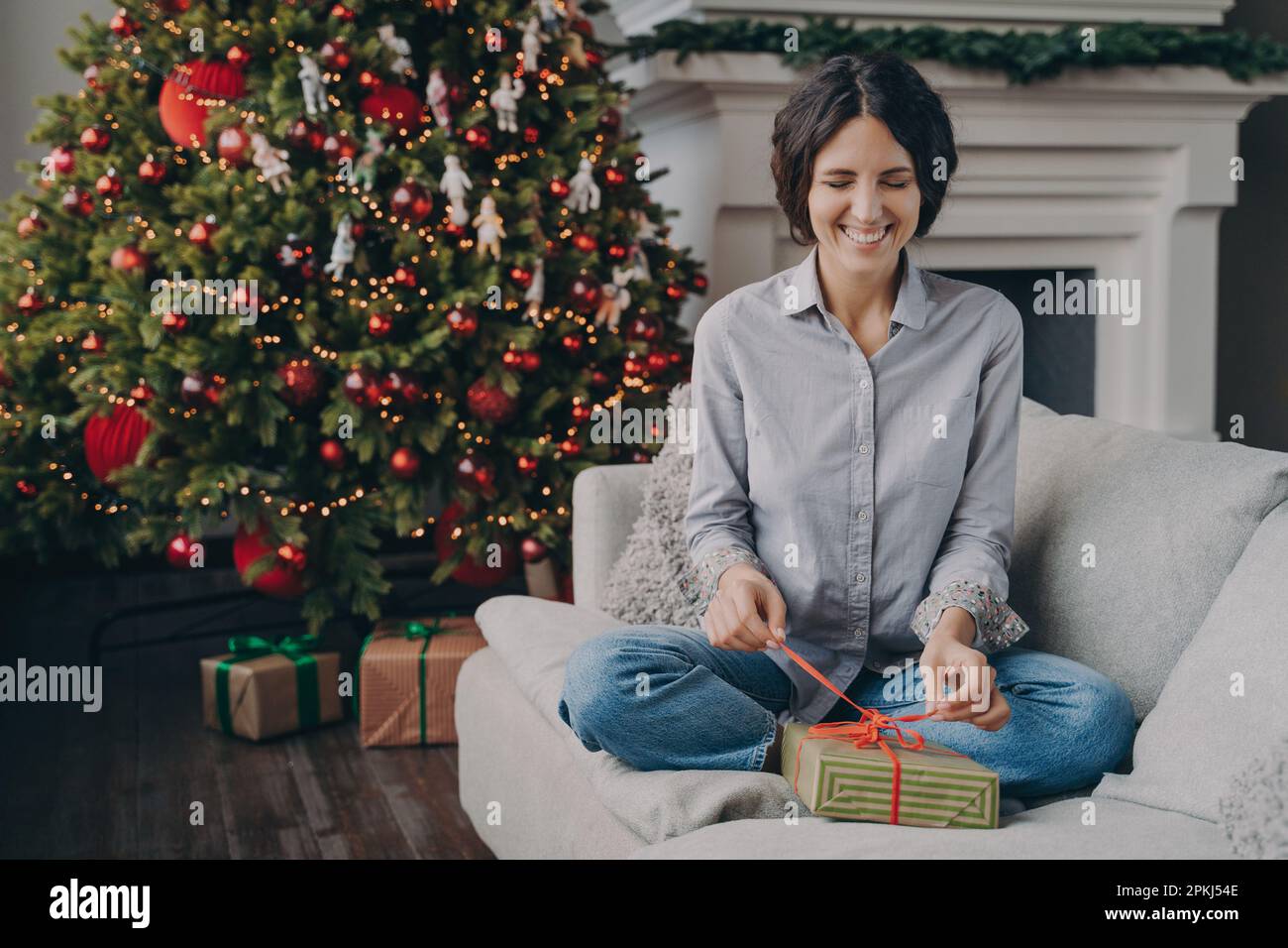 Eine junge, aufgeregte Italienerin lächelt verspielt mit geschlossenen Augen und versucht, ein Weihnachtsgeschenk auszupacken, indem sie das Band mit den Fingern hält, während sie auf dem bequemen Sofa sitzt Stockfoto
