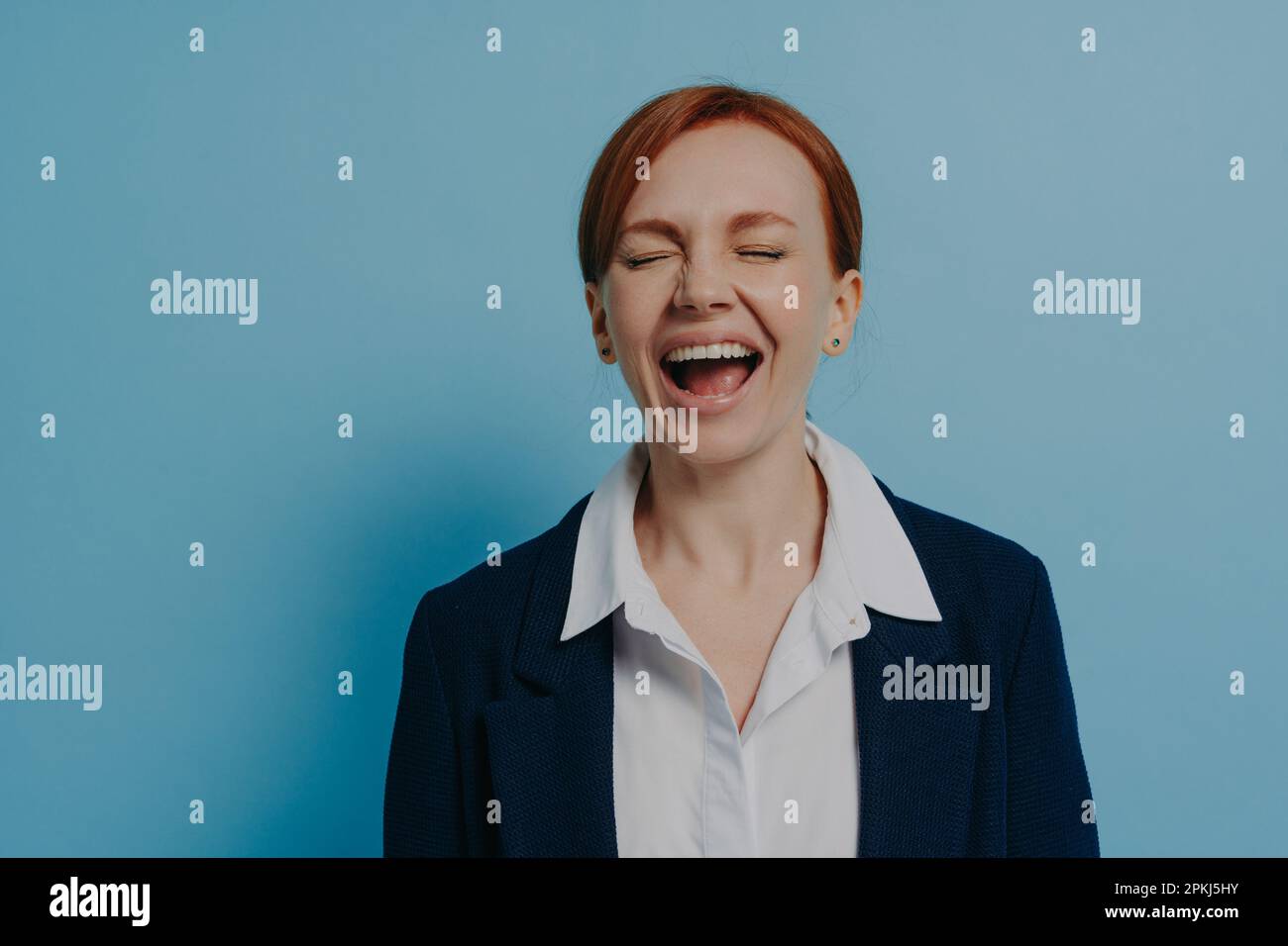 Studiofoto einer lachenden, glücklichen Frau in formeller Kleidung mit geschlossenen Augen, die isoliert an einer blauen Wand steht, einer Ingwer-Frau, die Spaß hat und lustige Anekdoten hört Stockfoto